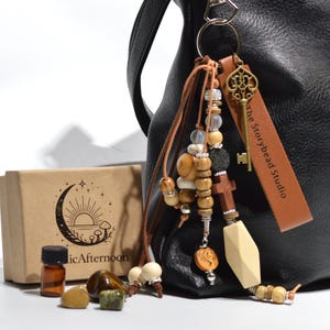Op de afbeelding: Een zwarte leren handtas met een decoratieve sleutelhanger. De sleutelhanger heeft houten kralen, een sleutelbedel en een leren label met de tekst "the Storybead Studio". Een kleine doos met een maan- en zonontwerp, een fles en edelstenen zijn ook aanwezig.