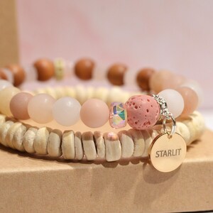 Puede incluir: Una pulsera apilada con cuentas de madera, cuentas de piedra rosa claro, blancas y color melocotón. Un pequeño dije redondo con la palabra "STARLIT" cuelga de la pulsera. La pulsera se exhibe en una caja de cartón.