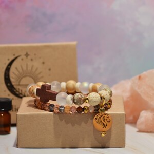 Pode incluir: Duas pulseiras de contas com uma cruz de madeira e um pingente de cavalo. As pulseiras est&atilde;o em uma pequena caixa marrom, com um pequeno frasco &acirc;mbar e uma caixa com um design de lua crescente e sol. Cristais de sal rosa no fundo.