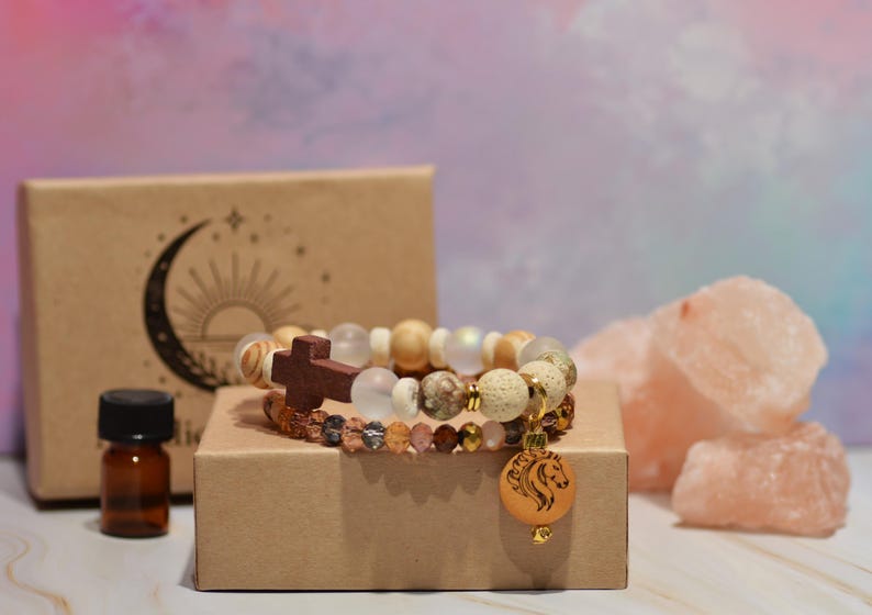 Pode incluir: Duas pulseiras de contas com uma cruz de madeira e um pingente de cavalo. As pulseiras est&atilde;o em uma pequena caixa marrom, com um pequeno frasco &acirc;mbar e uma caixa com um design de lua crescente e sol. Cristais de sal rosa no fundo.