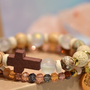 Pode incluir: Duas pulseiras empilhadas com tema religioso. A pulseira superior apresenta contas de madeira, contas transparentes e uma cruz marrom escuro. A pulseira inferior tem contas &acirc;mbar e pretas. Um pingente de madeira com um desenho de le&atilde;o pende da pulseira inferior.