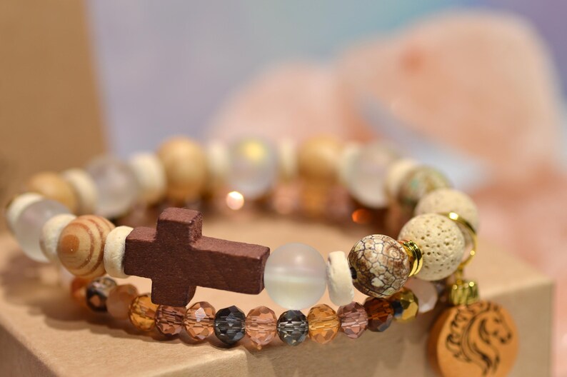 Pode incluir: Duas pulseiras empilhadas com tema religioso. A pulseira superior apresenta contas de madeira, contas transparentes e uma cruz marrom escuro. A pulseira inferior tem contas &acirc;mbar e pretas. Um pingente de madeira com um desenho de le&atilde;o pende da pulseira inferior.