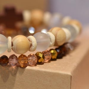 Pode incluir: Close-up de duas pulseiras de contas. A pulseira superior apresenta contas de madeira de cor clara e contas de vidro fosco. A pulseira inferior tem contas facetadas em tons de castanho, &acirc;mbar e dourado. As pulseiras est&atilde;o sobre uma superf&iacute;cie de cart&atilde;o.