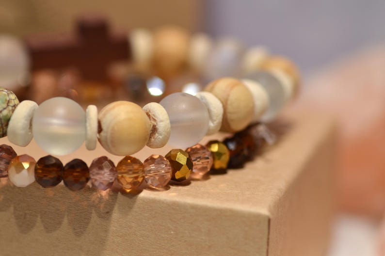 Pode incluir: Close-up de duas pulseiras de contas. A pulseira superior apresenta contas de madeira de cor clara e contas de vidro fosco. A pulseira inferior tem contas facetadas em tons de castanho, &acirc;mbar e dourado. As pulseiras est&atilde;o sobre uma superf&iacute;cie de cart&atilde;o.