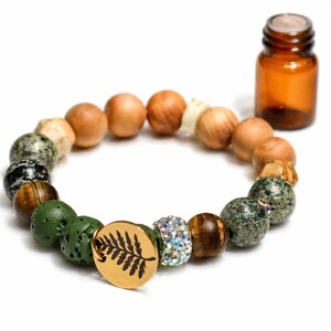 Op de afbeelding: Een kralenarmband met een verscheidenheid aan natuurlijke stenen en houten kralen, waaronder groene, bruine en gespikkelde stenen. Een kleine amberkleurige glazen fles staat in de buurt. De armband heeft een houten bedel met een varenontwerp.