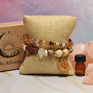 Pode incluir: Duas pulseiras de contas com contas castanhas, douradas e transparentes repousam sobre uma almofada de serapilheira. Um pequeno frasco de &oacute;leo essencial castanho com tampa preta fica ao lado de peda&ccedil;os de sal rosa do Himalaia. Uma caixa com o texto "NordicAfternoon" est&aacute; no fundo.