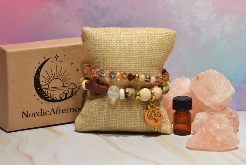Pode incluir: Duas pulseiras de contas com contas castanhas, douradas e transparentes repousam sobre uma almofada de serapilheira. Um pequeno frasco de &oacute;leo essencial castanho com tampa preta fica ao lado de peda&ccedil;os de sal rosa do Himalaia. Uma caixa com o texto "NordicAfternoon" est&aacute; no fundo.
