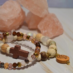 Pode incluir: Duas pulseiras de contas com uma cruz e um pingente de madeira. As pulseiras apresentam contas em tons de castanho, branco e transparente, com detalhes dourados. Rochas de sal rosa ao fundo.