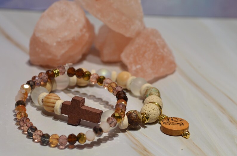 Pode incluir: Duas pulseiras de contas com uma cruz e um pingente de madeira. As pulseiras apresentam contas em tons de castanho, branco e transparente, com detalhes dourados. Rochas de sal rosa ao fundo.