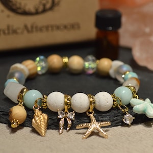 Può includere: Un braccialetto di perline con vari ciondoli, tra cui una conchiglia, una stella marina e un fiocco. Il braccialetto presenta perline azzurre, bianche e di legno, con dettagli dorati. Una piccola bottiglia e una scatola con la scritta "Nordic Afternoon" sono sullo sfondo.