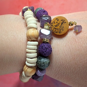 Op de afbeelding: Twee armbanden met kralen. Eén armband heeft licht houten kralen en natuurlijk gekleurde kralen. De andere armband heeft paarse, grijze en amethistkleurige kralen. Een houten bedel met een gesneden ontwerp en een kleine paarse kraal hangt aan de tweede armband.