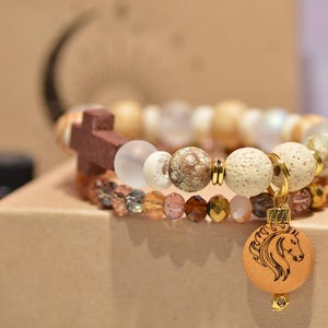 Pode incluir: Duas pulseiras de contas com uma cruz de madeira e um pingente de cavalo. As pulseiras apresentam uma variedade de contas em tons neutros, incluindo branco, marrom e transparente. O pingente de cavalo &eacute; redondo e tem um design detalhado da cabe&ccedil;a de cavalo.