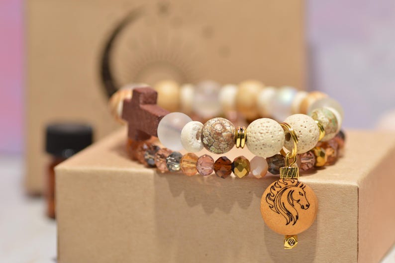 Pode incluir: Duas pulseiras de contas com uma cruz de madeira e um pingente de cavalo. As pulseiras apresentam uma variedade de contas em tons neutros, incluindo branco, marrom e transparente. O pingente de cavalo &eacute; redondo e tem um design detalhado da cabe&ccedil;a de cavalo.