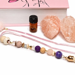 May include: A pink book, essential oil bottle, two pink Himalayan salt chunks, and a beaded bookmark with a tassel. The bookmark features various beads in shades of pink, purple, and gold. The items are arranged on a white surface.