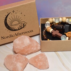 Pode incluir: Uma caixa de papel kraft com o texto "NordicAfternoon" e um logotipo de lua crescente, ao lado de uma caixa menor contendo uma pulseira de contas, uma garrafa pequena e uma cruz de madeira. Tr&ecirc;s pedras de sal rosa est&atilde;o em primeiro plano.