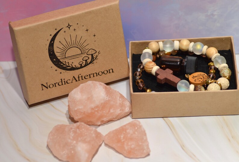Pode incluir: Uma caixa de papel kraft com o texto "NordicAfternoon" e um logotipo de lua crescente, ao lado de uma caixa menor contendo uma pulseira de contas, uma garrafa pequena e uma cruz de madeira. Tr&ecirc;s pedras de sal rosa est&atilde;o em primeiro plano.
