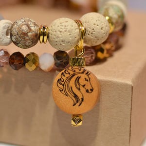 Pode incluir: Uma pulseira de contas com um pingente de cavalo. A pulseira apresenta uma variedade de contas em tons neutros, incluindo contas transparentes, castanhas e creme. Um pingente de madeira com um desenho de cabe&ccedil;a de cavalo pende.