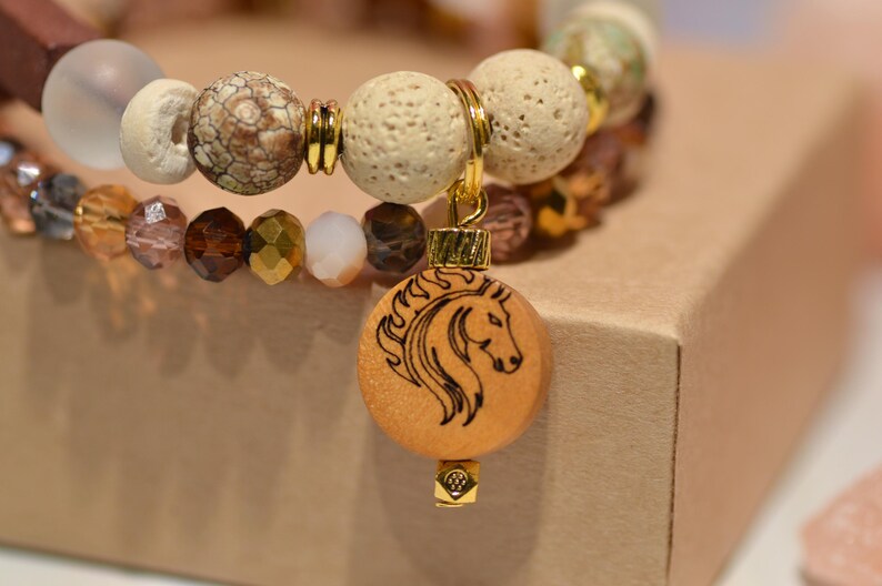 Pode incluir: Uma pulseira de contas com um pingente de cavalo. A pulseira apresenta uma variedade de contas em tons neutros, incluindo contas transparentes, castanhas e creme. Um pingente de madeira com um desenho de cabe&ccedil;a de cavalo pende.