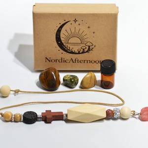 May include: A collection of natural stones, a small amber glass bottle, and a beaded pendant with a wooden cross and geometric shapes. A brown box with the text "NordicAfternoon" and a crescent moon design is in the background.