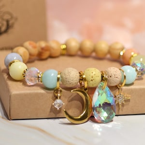 Może przedstawiać: Bransoletka z mieszanką jasnych koralików, w tym drewnianych, żółtych i niebieskich. Złote zawieszki to półksiężyc, gwiazda i kryształ w kształcie łzy. Bransoletka jest prezentowana na brązowym kartonowym pudełku.