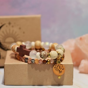 Pode incluir: Duas pulseiras de contas com uma cruz de madeira e um pingente de cavalo repousam sobre uma pequena caixa de papel&atilde;o. As pulseiras apresentam v&aacute;rias contas em tons terrosos. Uma pequena garrafa &acirc;mbar e uma caixa com um desenho de lua crescente e sol tamb&eacute;m est&atilde;o presentes.