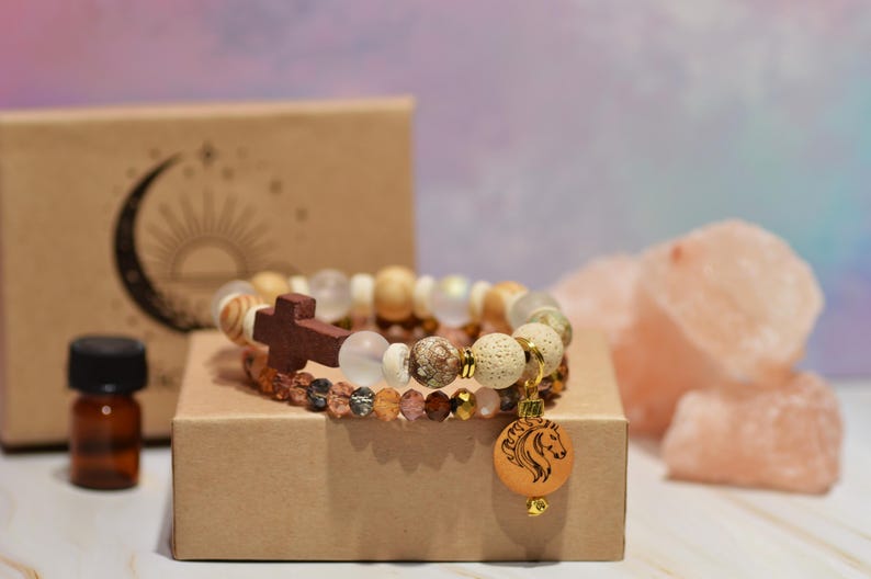 Pode incluir: Duas pulseiras de contas com uma cruz de madeira e um pingente de cavalo repousam sobre uma pequena caixa de papel&atilde;o. As pulseiras apresentam v&aacute;rias contas em tons terrosos. Uma pequena garrafa &acirc;mbar e uma caixa com um desenho de lua crescente e sol tamb&eacute;m est&atilde;o presentes.