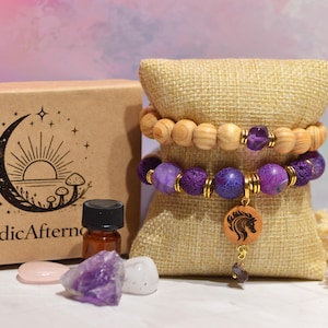 Peut inclure: Deux bracelets de perles avec des perles en bois et violettes sont présentés sur un présentoir en toile de jute. Une petite bouteille, des cristaux et une boîte avec le texte "NordicAfternoon" et un logo de croissant de lune sont également présents. Les bracelets ont des accents dorés et un charme de cheval.