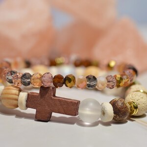 Pode incluir: Duas pulseiras de contas, uma com um pingente de cruz de madeira. As pulseiras apresentam uma mistura de contas marrons, douradas e transparentes, e contas de madeira e brancas. O fundo est&aacute; desfocado.