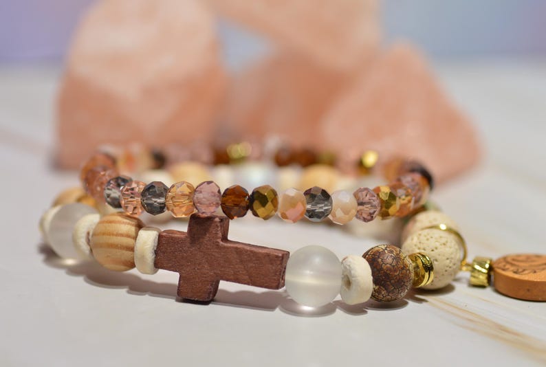 Pode incluir: Duas pulseiras de contas, uma com um pingente de cruz de madeira. As pulseiras apresentam uma mistura de contas marrons, douradas e transparentes, e contas de madeira e brancas. O fundo est&aacute; desfocado.