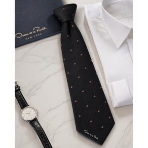 May include: A black necktie featuring a pattern of small red and white cherry motifs, with the text Oscar de la Renta printed in white near the bottom tip. The tie is displayed alongside a white dress shirt, a wristwatch, and a dark blue box.