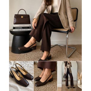 May include: Brown slingback heels with a textured pattern. The heels have a block heel and a strap that goes around the ankle. The shoes are paired with brown pants and a beige blazer. A brown handbag and books are also in the image.