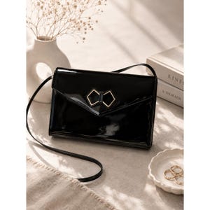 May include: A black patent leather envelope-style crossbody bag with a thin shoulder strap. The front flap features a geometric gold-tone metal bow ornament. The bag has a glossy, reflective finish and a structured rectangular shape.
