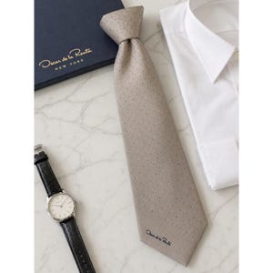 May include: A beige dotted tie with the "Oscar de la Renta NEW YORK" logo, a white dress shirt, and a silver watch with a black leather strap. The tie is knotted on a marble surface.
