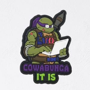 May include: A patch featuring a green cartoon turtle wearing a purple mask and glasses, holding a laptop. The patch has the words "COWABUNGA IT IS" in purple and green. The turtle is wearing a brown vest and blue straps.
