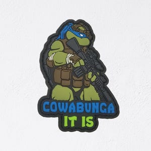 May include: A patch featuring a green turtle-like character with a blue mask, holding a rifle. The patch has the words "COWABUNGA IT IS" in blue and green lettering.