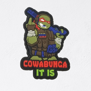 May include: A cartoon patch featuring a green turtle-like character in tactical gear, holding a blue weapon. The character is wearing a red mask and headset. The text "COWABUNGA IT IS" is displayed in red and green.