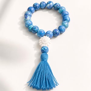May include: A circular beaded bracelet featuring round beads in various shades of blue with a marbled pattern. A single white, textured, star-shaped bead is positioned near the bottom, followed by one blue bead and a long, blue fabric tassel.