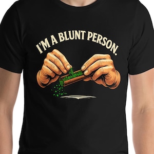 May include: Black t-shirt featuring the text "I'M A BLUNT PERSON." The graphic depicts hands rolling a blunt with green herbs. The shirt is made of a soft material and has short sleeves.