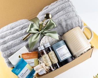 Puede incluir: Una caja de regalo que contiene una manta de felpa gris doblada, una taza de cerámica acanalada de color crema, una lata de té English Breakfast, dos tubos de vidrio con frutos secos cubiertos de chocolate, un tarro de miel y dos barras de chocolate, todo atado con una cinta verde salvia.