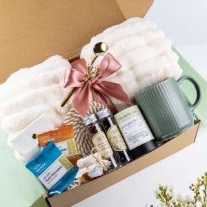 Puede incluir: Una caja de regalo que contiene una manta de piel sint&eacute;tica color crema atada con una cinta rosa empolvado, una taza de cer&aacute;mica acanalada verde salvia, t&eacute; verde al jazm&iacute;n Tea With Tae, dos viales de almendras cubiertas de chocolate y chocolate Verse.