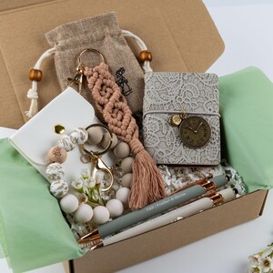 Puede incluir: Una caja de regalo que contiene un llavero de macram&eacute; beige con borla, una peque&ntilde;a bolsa de arpillera, un tarjetero blanco, un llavero de cuentas con detalles dorados, un cuaderno gris con encaje y dije de reloj, y dos bol&iacute;grafos Feather & Bloom.