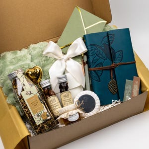 Puede incluir: Una caja de regalo que contiene un diario azul cerceta con un dije de hoja, una manta de felpa verde salvia, un lazo de sat&eacute;n blanco, una botella de t&eacute; de albaricoque y jazm&iacute;n, almendras cubiertas de chocolate, miel y un sobre verde.
