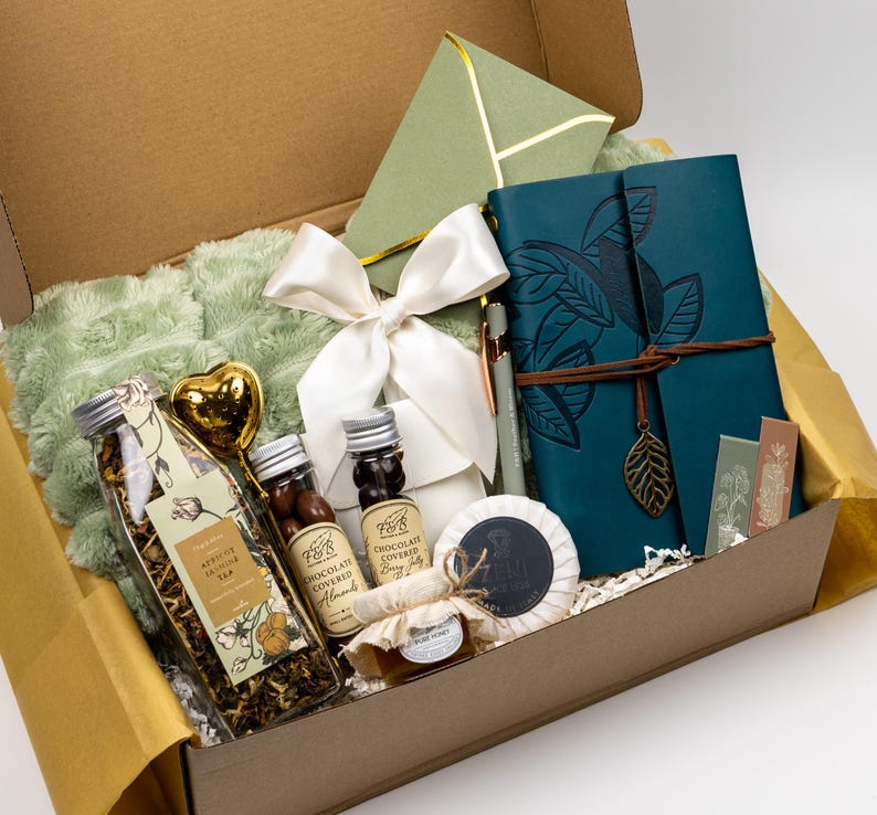 Puede incluir: Una caja de regalo que contiene un diario azul cerceta con un dije de hoja, una manta de felpa verde salvia, un lazo de sat&eacute;n blanco, una botella de t&eacute; de albaricoque y jazm&iacute;n, almendras cubiertas de chocolate, miel y un sobre verde.