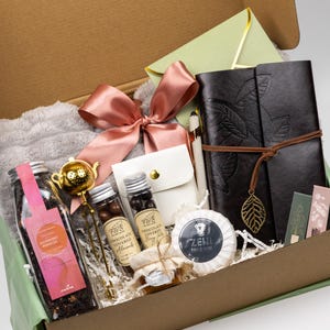 Puede incluir: Una caja de regalo que contiene un diario marr&oacute;n oscuro con un dije de hoja, un infusor de t&eacute; dorado, un frasco de t&eacute; de hibisco y frambuesa, dos frascos peque&ntilde;os de snacks cubiertos de chocolate, una bolsa blanca, un jab&oacute;n Zeni, un lazo rosa y un sobre verde claro.