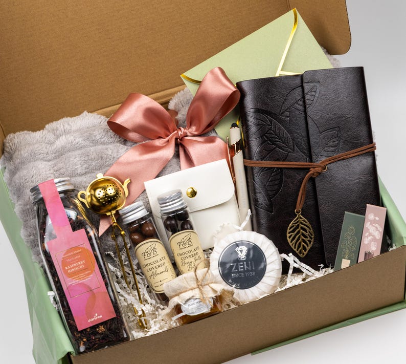 Puede incluir: Una caja de regalo que contiene un diario marr&oacute;n oscuro con un dije de hoja, un infusor de t&eacute; dorado, un frasco de t&eacute; de hibisco y frambuesa, dos frascos peque&ntilde;os de snacks cubiertos de chocolate, una bolsa blanca, un jab&oacute;n Zeni, un lazo rosa y un sobre verde claro.