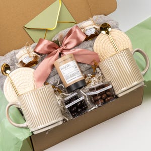 Puede incluir: Una caja de regalo con dos tazas beige acanaladas con cucharas doradas, dos posavasos redondos trenzados, una manta gris de felpa, una cinta de sat&eacute;n rosa, un bote TEA WITH TAE, dos tarros de miel y dos cajas de frutos secos con chocolate.