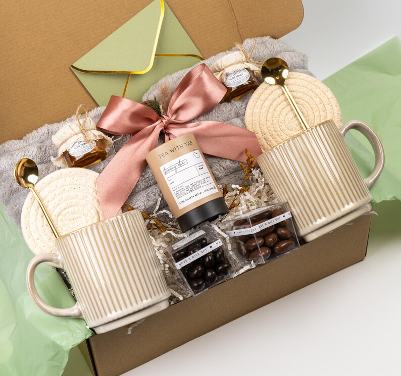 Puede incluir: Una caja de regalo con dos tazas beige acanaladas con cucharas doradas, dos posavasos redondos trenzados, una manta gris de felpa, una cinta de sat&eacute;n rosa, un bote TEA WITH TAE, dos tarros de miel y dos cajas de frutos secos con chocolate.