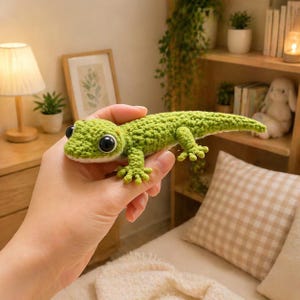 May include: A handcrafted, green crochet gecko with large black eyes. The reptile features detailed tiny feet and a long tail. The gecko is held in a person's hand, highlighting the texture and craftsmanship of the crochet work.