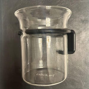 Könnte beinhalten: Eine transparente Bodum-Kaffeepresse aus Glas mit schwarzem Griff und Band. Die Glaskaraffe hat einen ausgestellten oberen Teil und einen zylindrischen Körper. Das Bodum-Logo ist auf die Seite des Glases gedruckt.