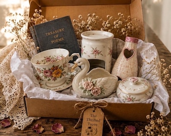 May include: A gift box containing a Treasury of Poems book, a floral teacup and saucer, a swan-shaped dish with pink roses, a ceramic wax warmer, a heart-cutout speckled vase, and a lidded floral jar, all nestled in white tissue paper with dried flowers.